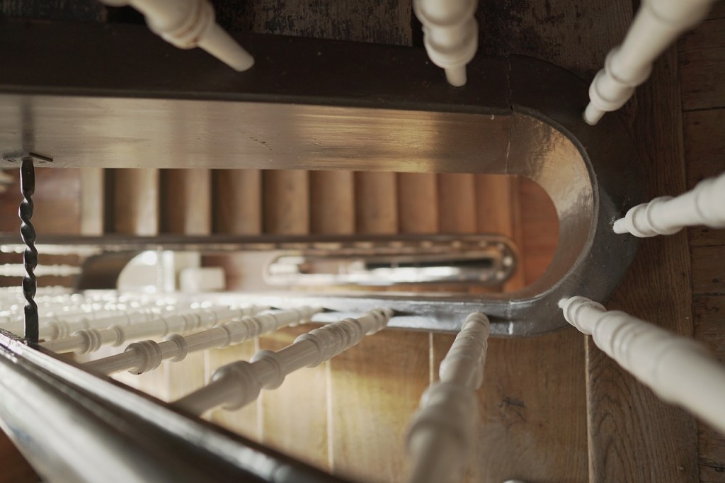 Ein Treppenhaus in Holzoptik, ein Anblick aus einem oberen Geschoss, hinunter zwischen den Treppengeländern. 