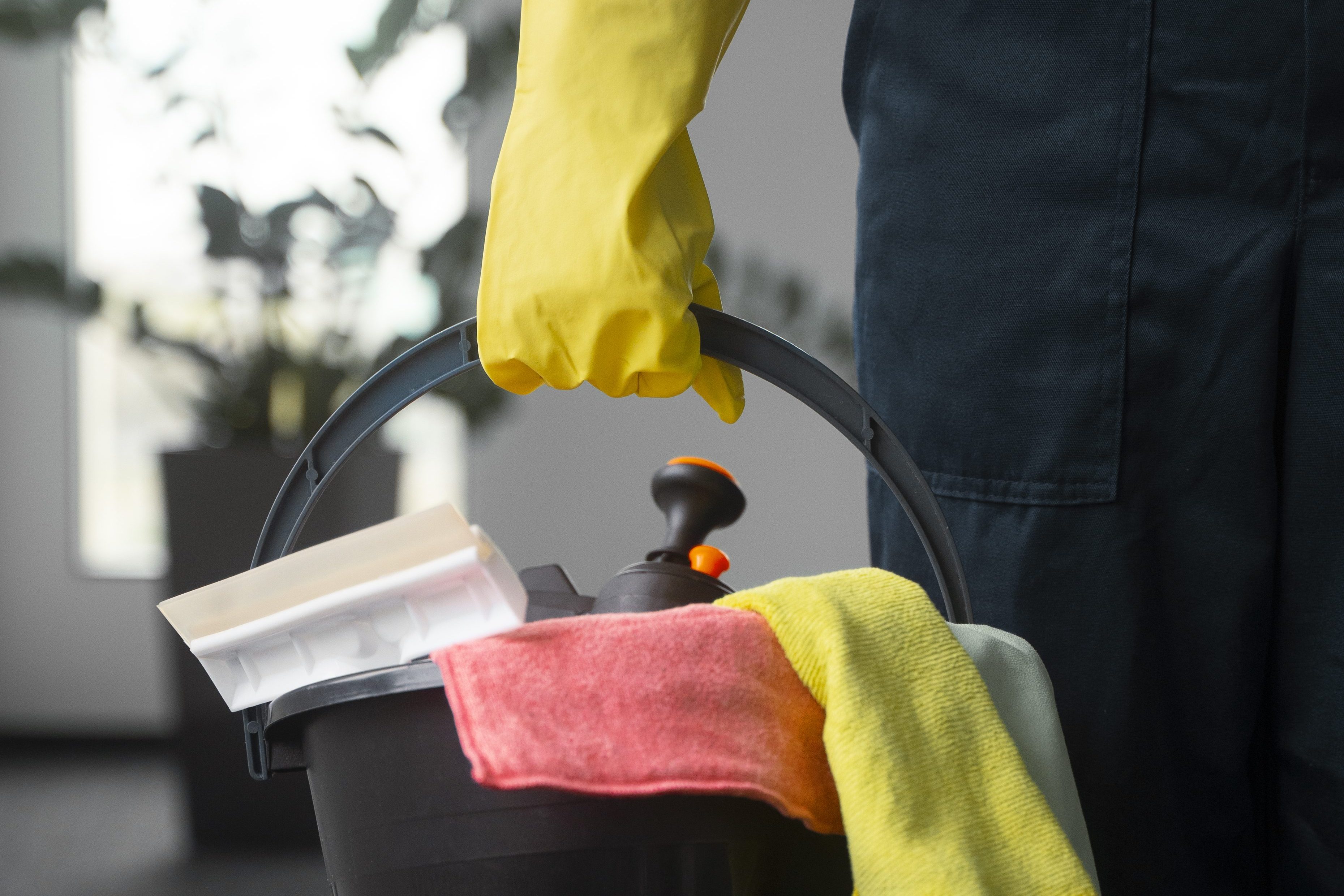 Hand mit gelben Handschuhe, hält einen Eimer voll mit Reinigungstüchern und Reinigungsmitteln, Motiv für Gebäudereinigung